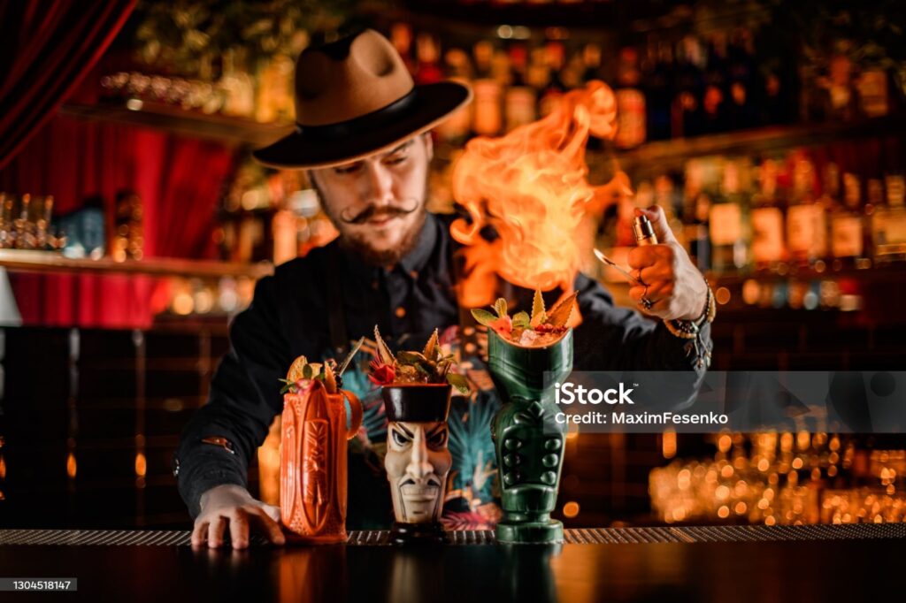 Barman care prepară cocktailuri cu flăcări spectaculoase în fața unui bar, utilizând un spray pentru a aprinde băutura.